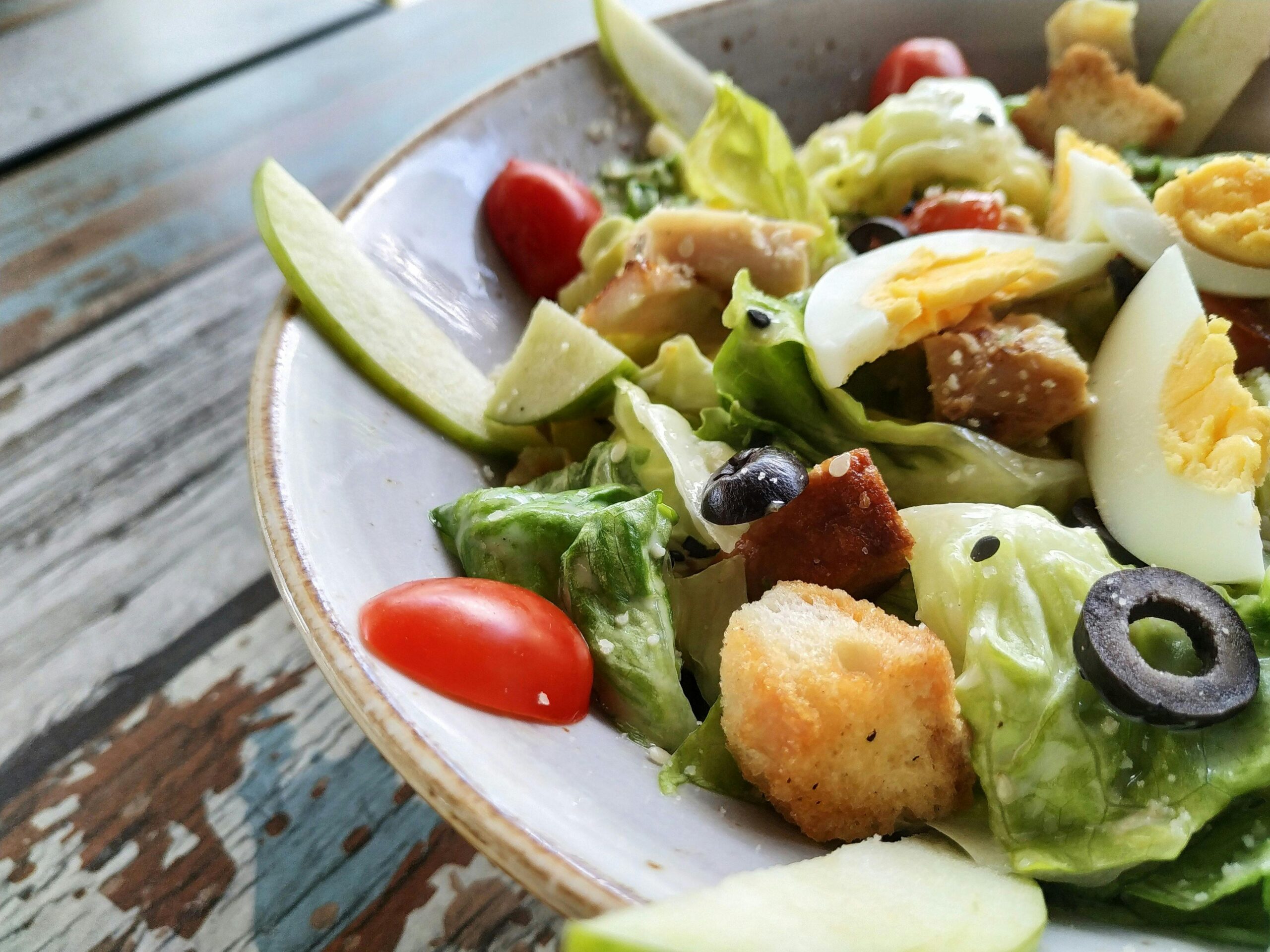 A close-up of a classic Caesar salad with crisp lettuce, boiled eggs, crunchy croutons, and olives, served fresh on a rustic table — a timeless favorite at top Italian restaurants near me?.